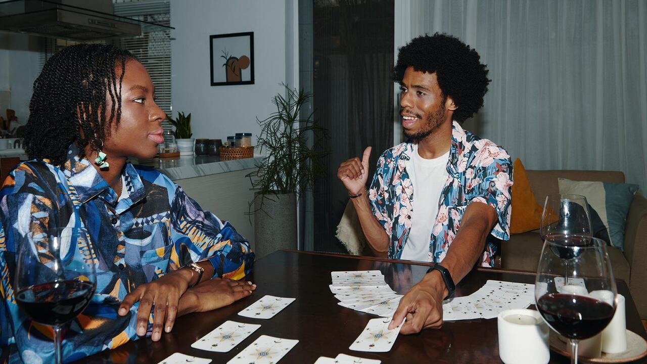 Two individuals seated at a dining table with candles, wine glasses, and tarot cards spread out.