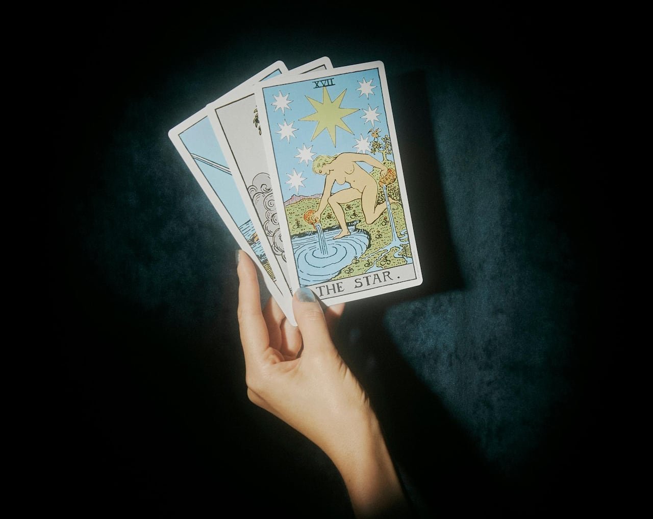A hand displaying three tarot cards against a dark blue velvet backdrop.